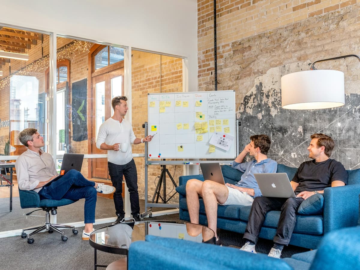 Team members with laptops in a meeting room as a colleague presents information at a whiteboard