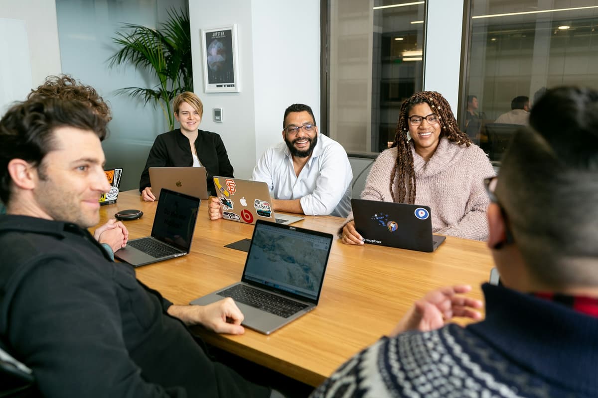 Four professionals seated with laptops in a board meeting, listening to a presenter
